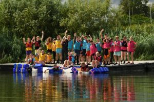Juniors in yellow, blue and pink team bibs waving from a jetty preparing to launch home-made rafts