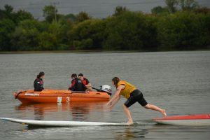 A yellow team member running the gauntlet of floating SUP and windsurf boards, with the safety boat behind